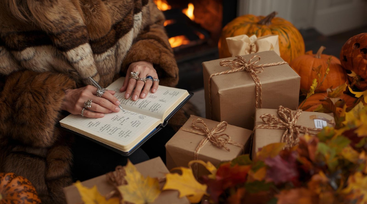 A person wearing a fur coat writes in an open notebook while sitting near a cozy fireplace, surrounded by pumpkins, wrapped gifts tied with twine, and colorful autumn leaves.
